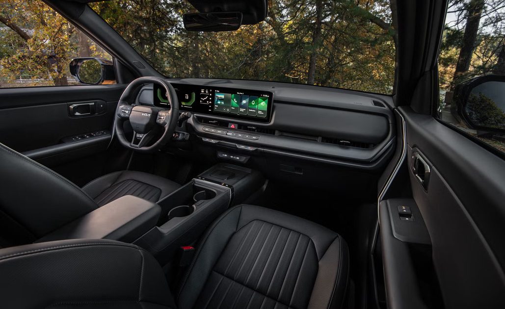 vehicle interior view, digital display and center console, black seats, wooded landscape visible through side windows.