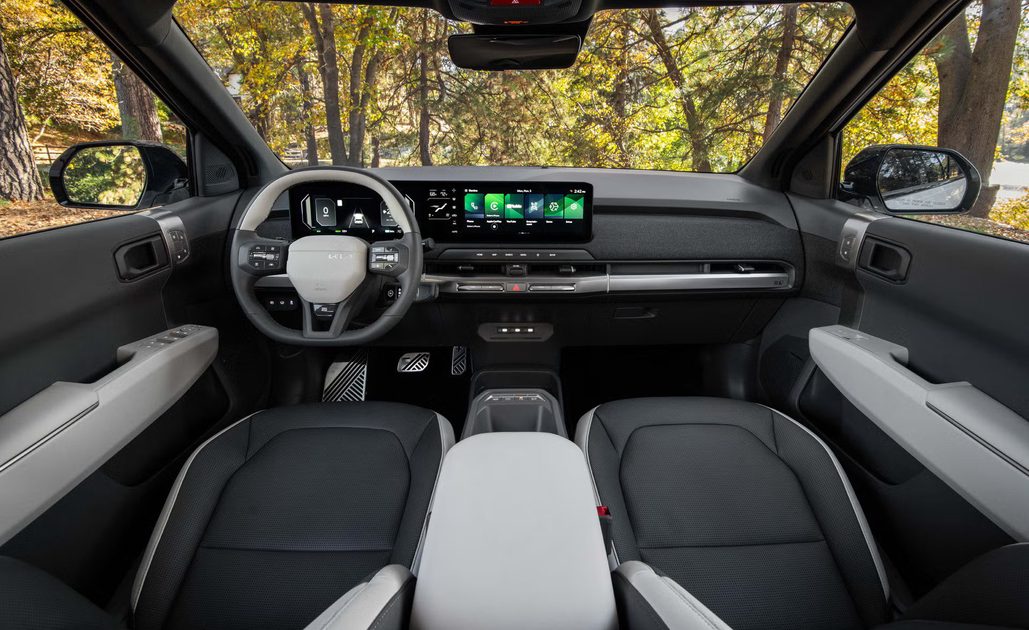vehicle interior view, dashboard with dual screens and steering wheel, front seats and forest visible through windshield.